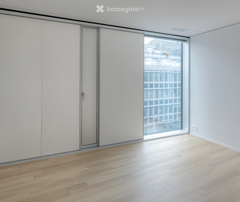 Empty room with light wooden flooring, white walls, large window with a view of a modern building, and white sliding closet doors.