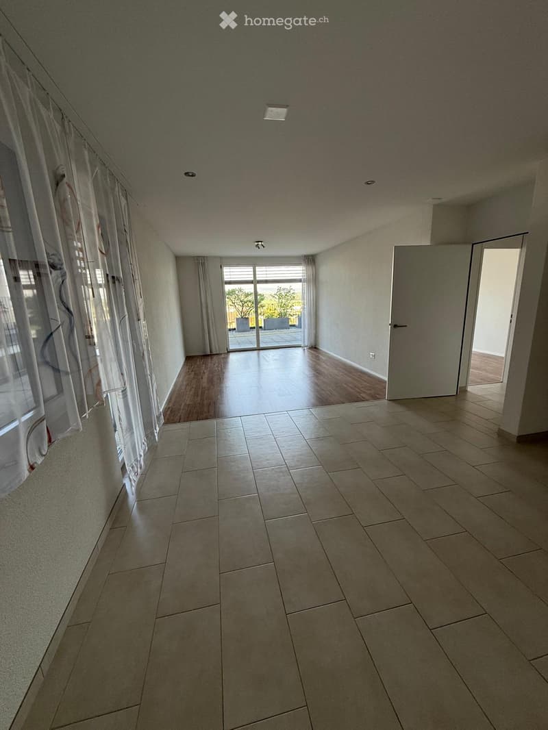 Empty room with tiled floor, wooden flooring in the adjacent area, large windows with curtains, and a sliding glass door leading to an outdoor space.