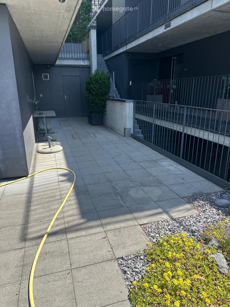 Concrete patio area with a small garden bed featuring yellow flowers, a metal railing, and a small table with a chair.