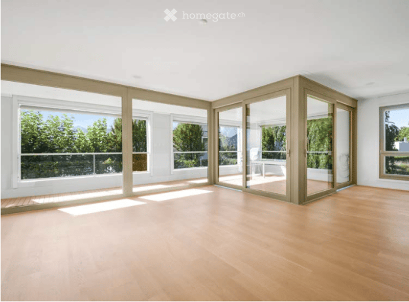 Bright, empty room with large windows and sliding glass doors leading to a balcony, featuring a wooden floor and white walls.