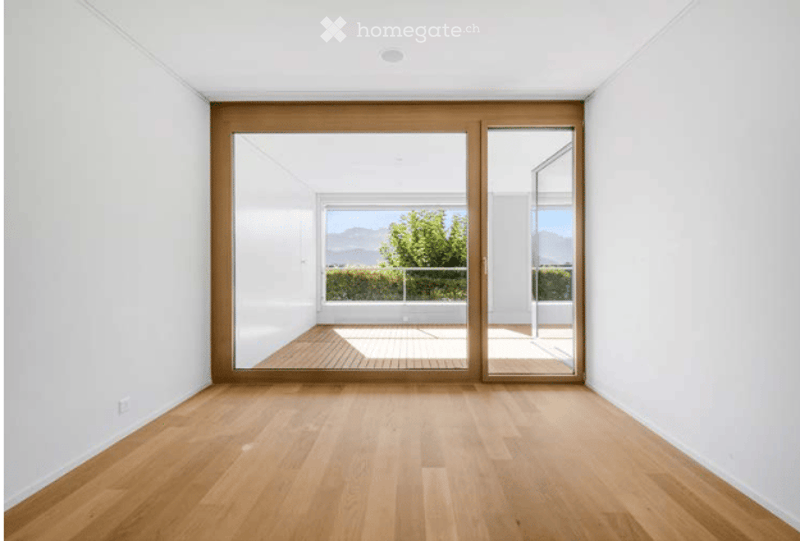 Bright, unfurnished room with light wooden flooring and large glass sliding doors leading to a balcony with a scenic mountain view.