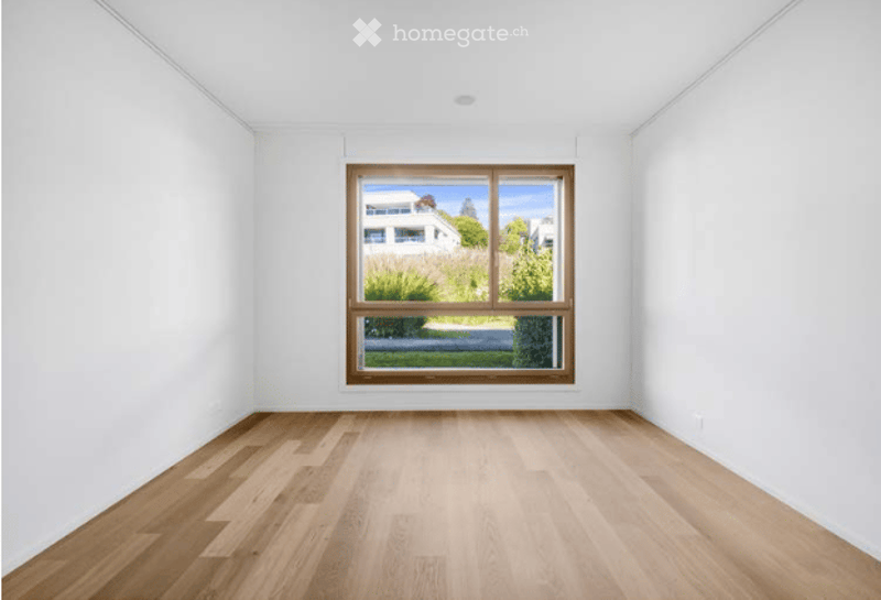 Empty room with white walls, wooden floor, and large window showing outdoor view.