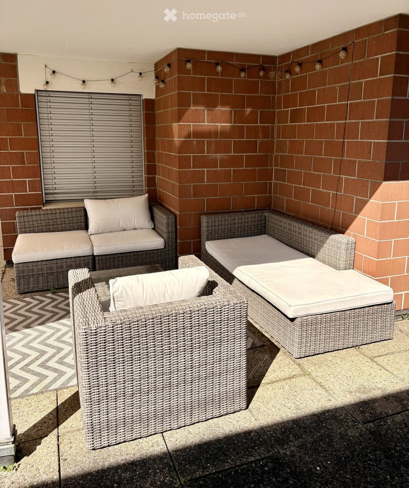 A cozy outdoor seating area with wicker furniture, including a sofa, chairs, and an ottoman, set against a brick wall with string lights.