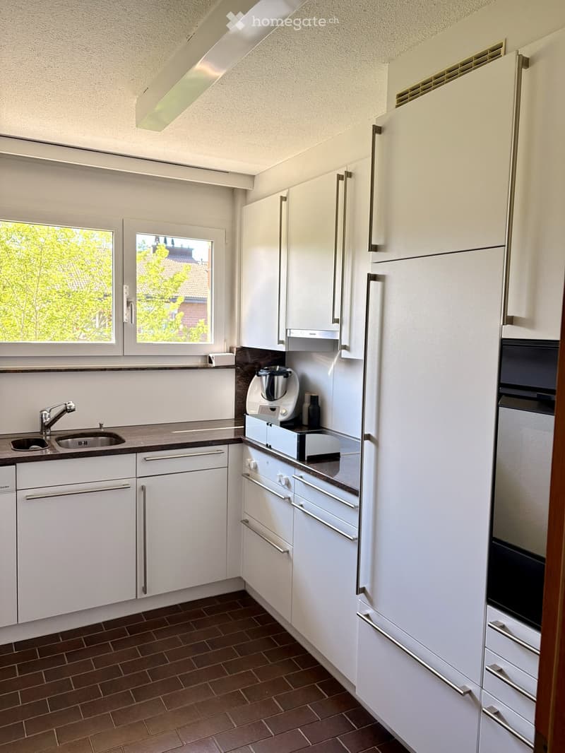 Modern kitchen with white cabinets, stainless steel appliances, and a dark brown tiled floor. Features include a double sink, a blender on the counter, and a refrigerator with a built-in oven.