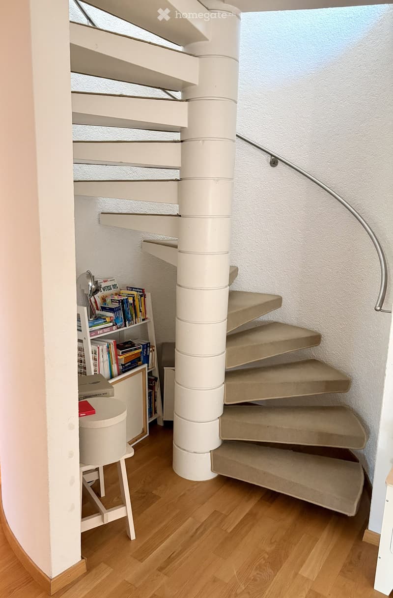 Spiral staircase with beige carpeted steps and white central column, adjacent to a small bookshelf filled with books and a small white stool.