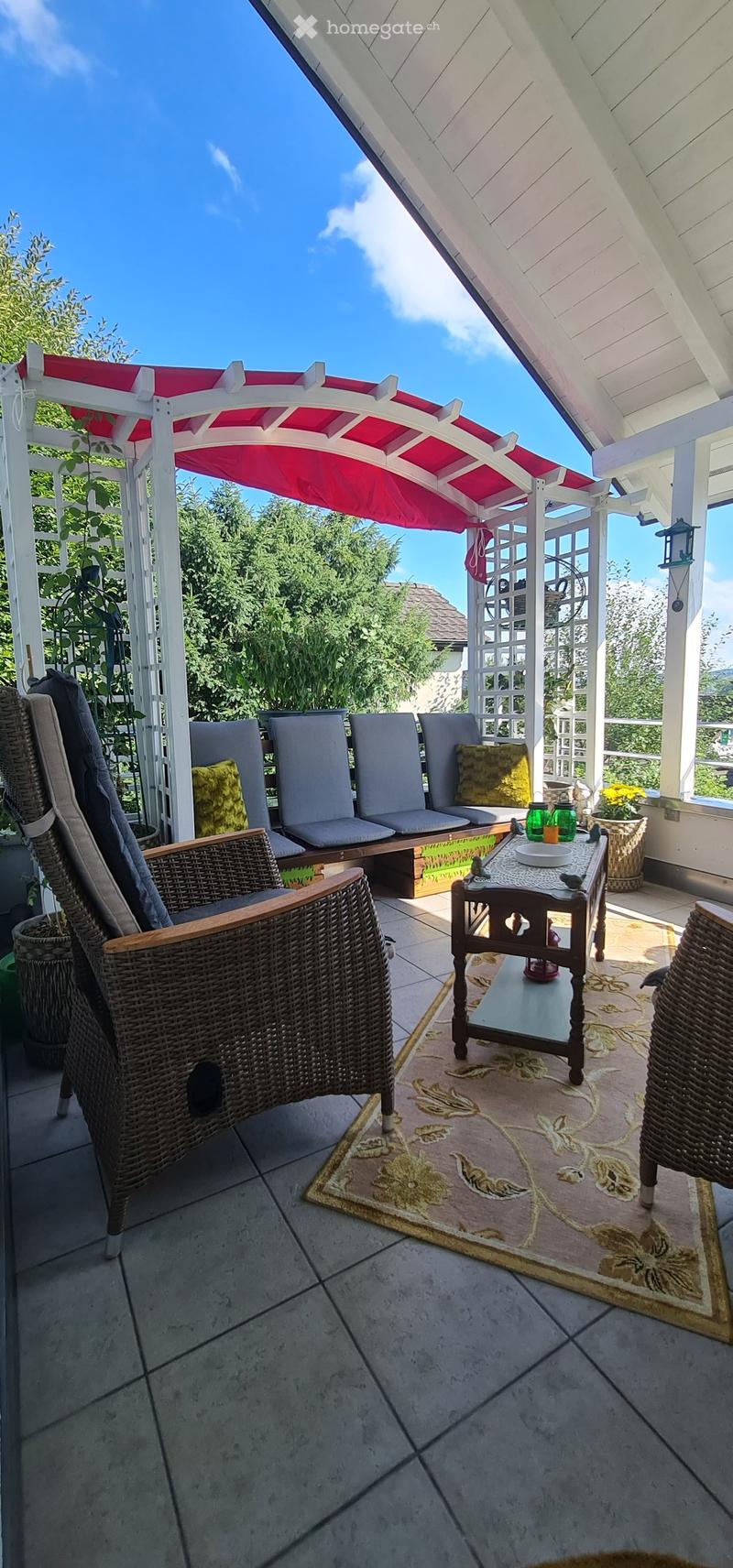 A covered patio with a tiled floor, a wicker chair, a small table with cups, and potted plants.