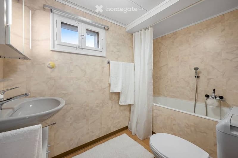 Bathroom with a sink, bathtub, toilet, and white towels. Features a window with a curtain and a small white rug on the floor.