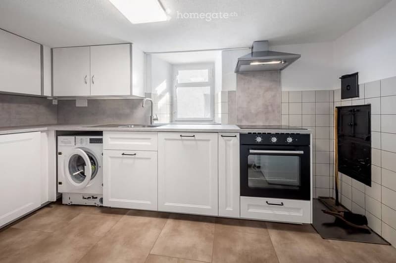 Modern kitchen with white cabinets, a sink, an oven, and a washing machine. The space features wooden flooring and a window above the sink.