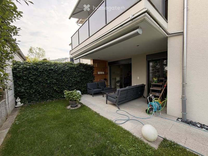 Well-maintained garden with a small lawn, a patio area with seating, a small table, and a potted plant. The patio is connected to a covered outdoor space with additional seating and a small kitchen area.