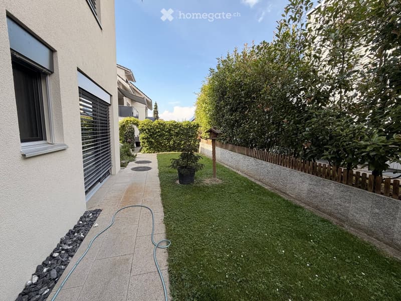Small, well-maintained garden with a paved pathway, grassy area, small wooden fence, and potted plant. The garden is bordered by a house with large windows and a raised ground floor.