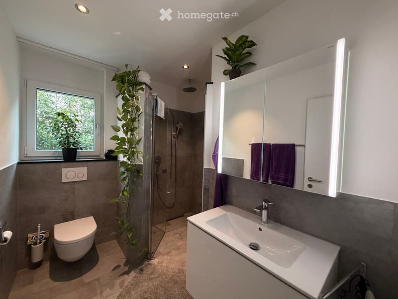 Modern bathroom with a large sink, toilet, shower, and a large mirror with built-in lighting. Features include a window with a plant, a towel rack with purple towels, and a sleek, contemporary design.