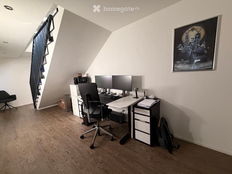 Modern, well-lit attic office with wooden flooring, a sleek black office chair, a white desk, dual monitors, and a large framed poster on the wall.
