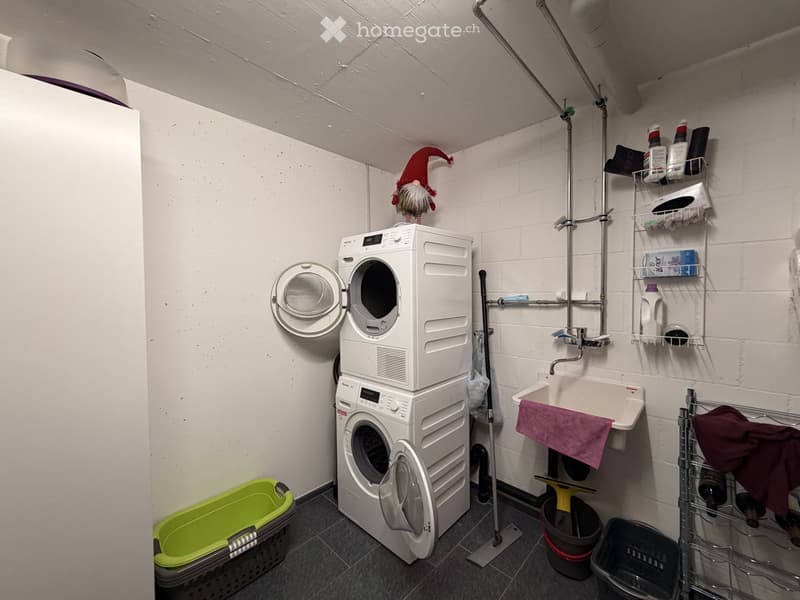 Compact bathroom with a stacked washing machine and dishwasher unit, a sink with a towel, shelves holding cleaning supplies, and various laundry baskets.