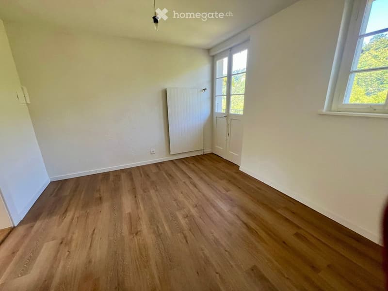 Empty room with light wooden flooring, white walls, and large windows allowing natural light. Features a white radiator and a door leading to another space.