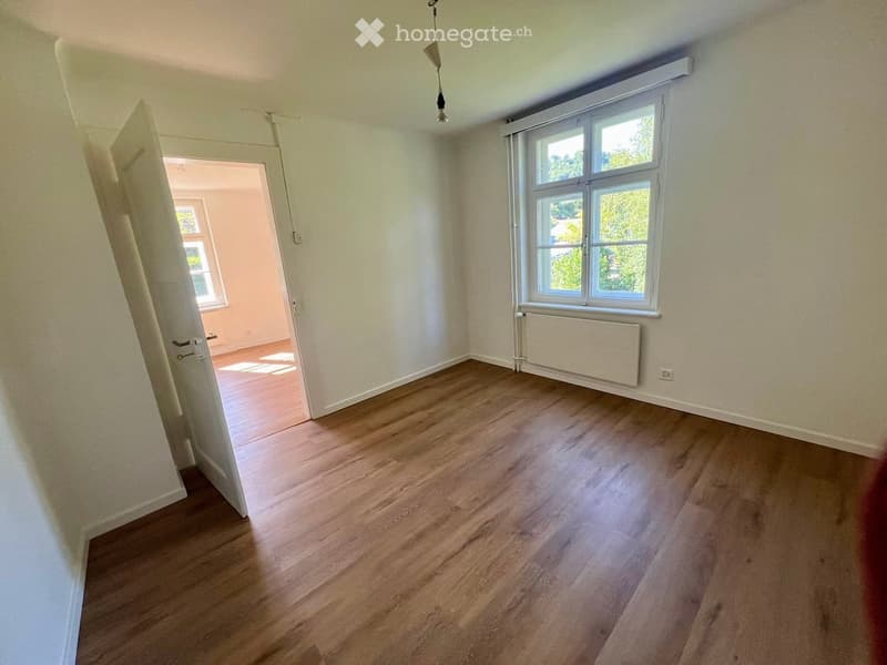 Empty room with light-colored wooden flooring, white walls, and a large window with white frames. A radiator is visible below the window, and a doorway leads to another room with similar flooring.