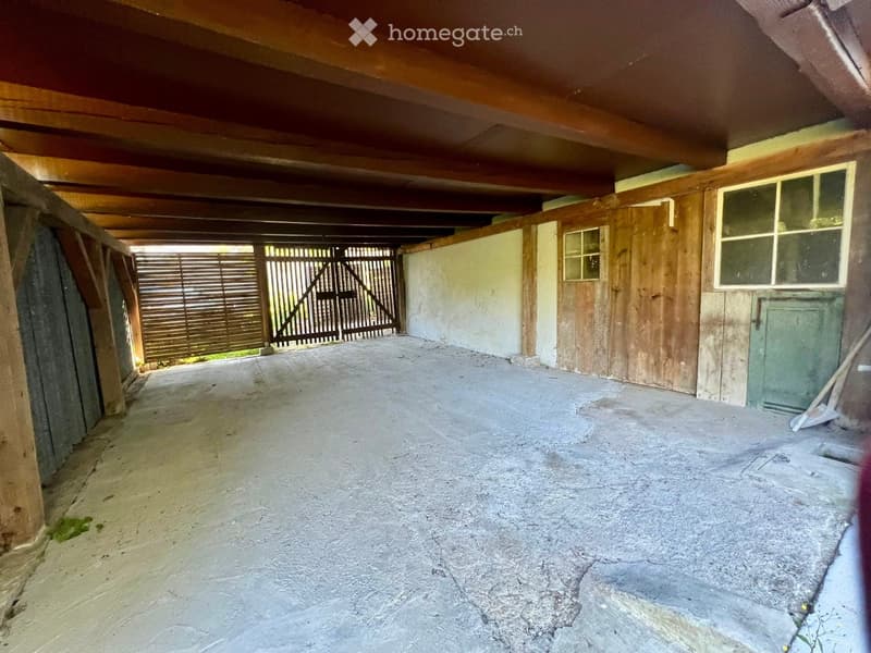 Empty garage with wooden beams, concrete floor, wooden doors, and a metal gate leading outside.