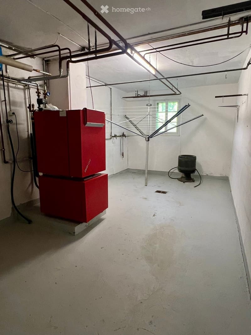 Empty utility room with concrete floor, white walls, exposed pipes, and a small window. Contains a red water heater unit and a clothes drying rack.