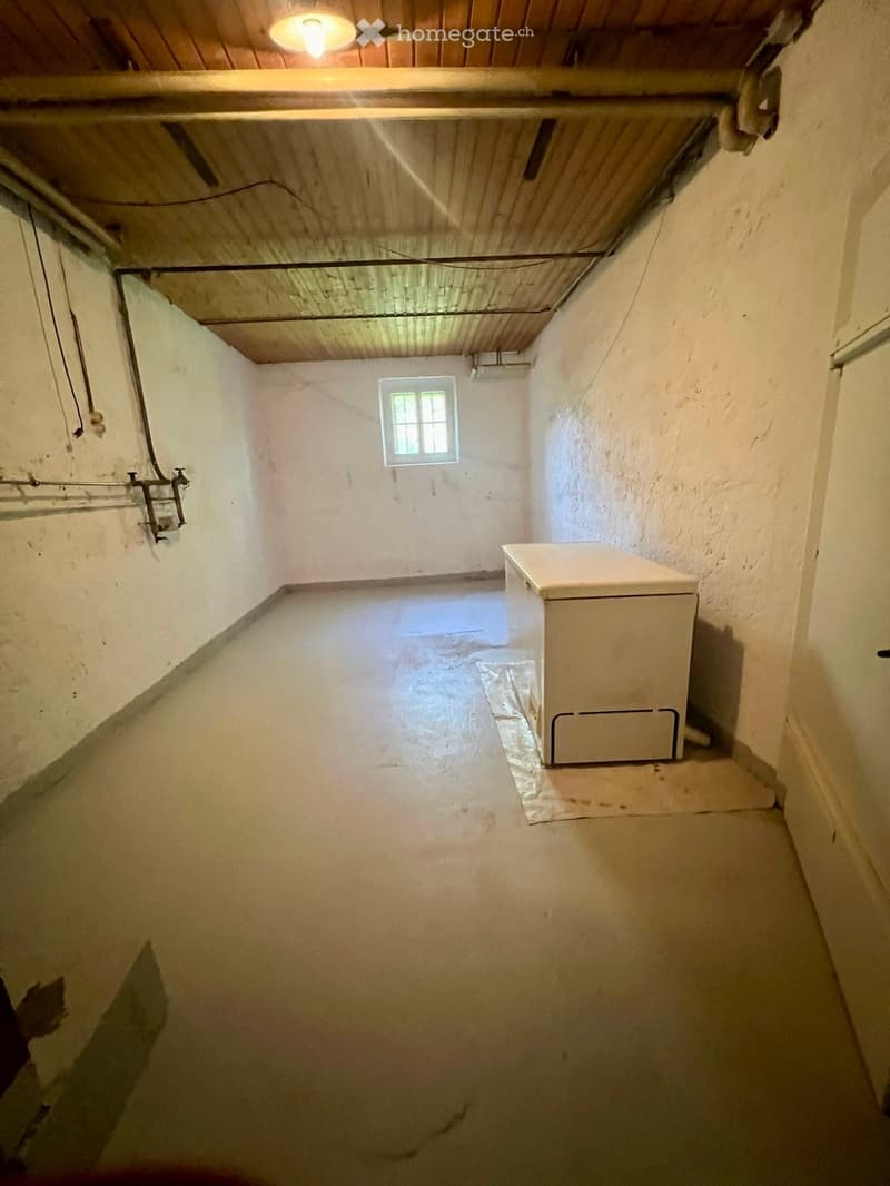 Empty room with white walls, a small window, and a single appliance placed against the wall. The ceiling has exposed wooden beams and electrical wiring.