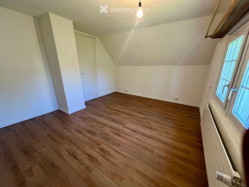 Empty attic room with wooden flooring, white walls, and a single window letting in natural light. The room features a slanted ceiling and a closed door leading to another area.