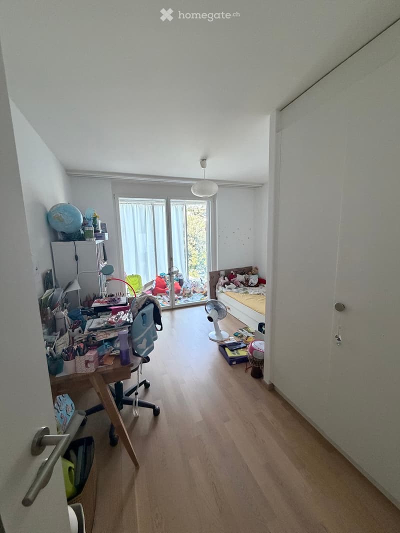 A multi-functional room with a desk area, a bed, and a fan. The room features wooden flooring, large windows with curtains, and various personal items including a globe, books, and toys.