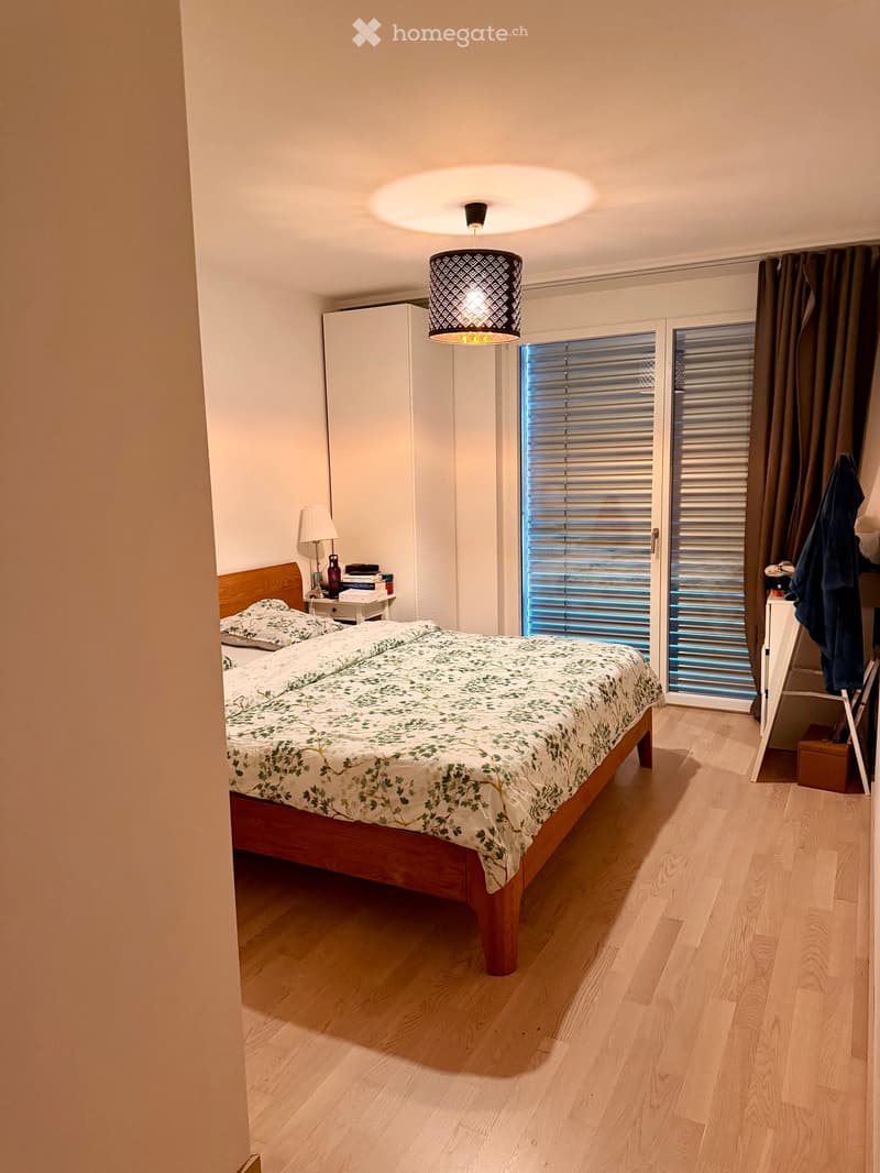 Modern bedroom with wooden flooring, a double bed with a leafy duvet, a nightstand with a lamp, books, and a wardrobe. A large window with blinds and a pendant light fixture is visible.