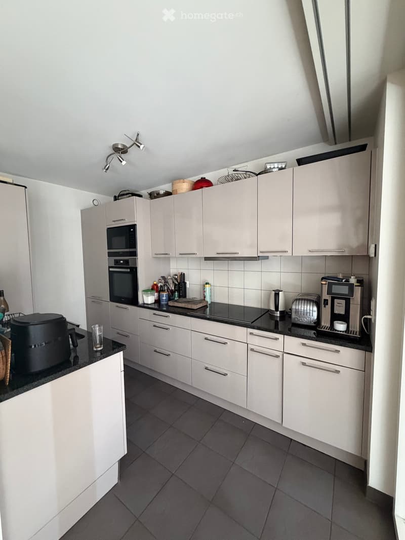 Modern kitchen with white cabinets, black countertops, and various appliances including a microwave, oven, coffee maker, and toaster. The kitchen features a dark tiled floor and a sleek, contemporary design.