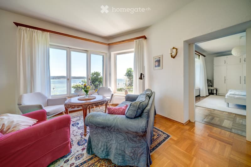 Modern living room with a view of the sea. The room has white walls, wooden floor, white curtains, and a wooden lamp