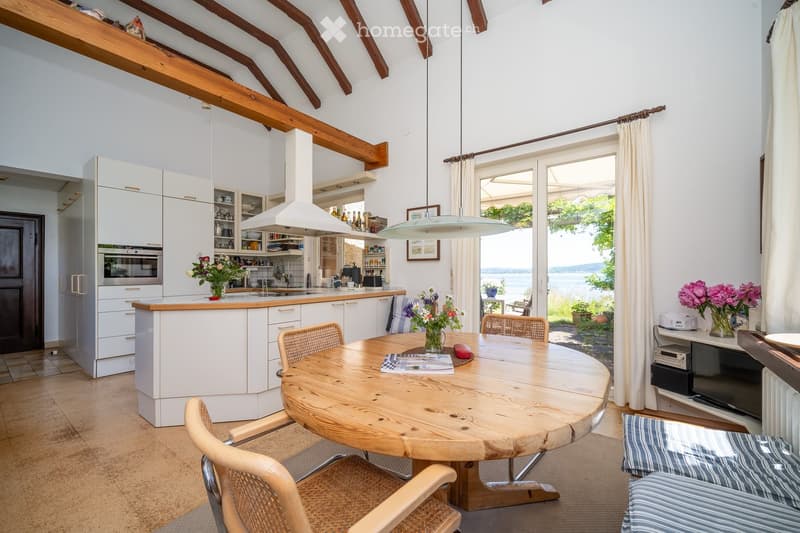 Wooden round table, armchairs, wooden kitchen island, white cabinetry, overhead lighting, sliding doors, view of the water