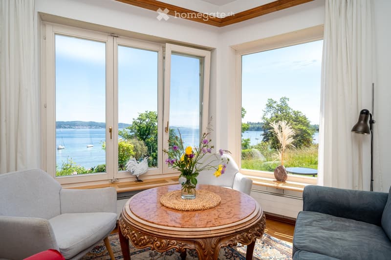 furnished room with couch, chairs, table, vases with flowers, view to the lake, windows with white curtains