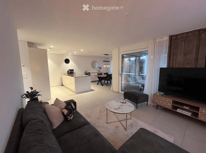 Modern open-plan living and dining area with a grey sofa, coffee table, ottoman, TV stand, kitchen island, dining table, chairs, and a potted plant. Large sliding glass doors lead to a balcony.
