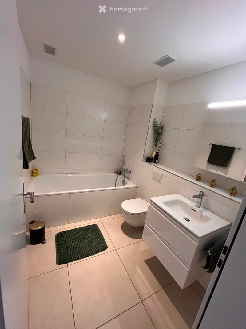 Modern bathroom with white tiles, bathtub, toilet, sink, mirror, towel, and a small green rug on the floor.