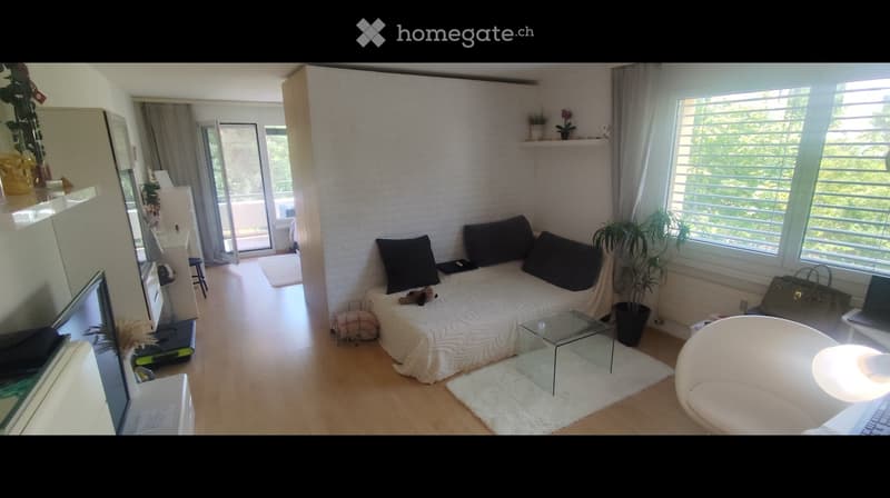 A modern, well-lit living room with a white brick accent wall, a sofa with black cushions, a glass coffee table, a white armchair, a laptop on a small table, a mirror, and a potted plant near the window. The room features large windows with blinds and a s