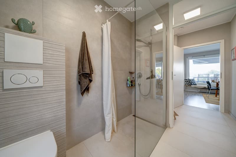 Modern bathroom with a glass-enclosed shower, toilet, towel, and decorative item on a shelf. The shower features a modern faucet and a white curtain. The bathroom has light-colored tiles and a sleek design.