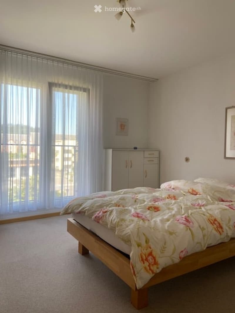 Bright bedroom with a wooden bed, floral duvet, white curtains, and a built-in wardrobe.