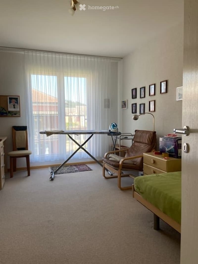 A well-lit bedroom with a bed, a wooden chair, an ironing board, and a lamp. The room features large windows with sheer curtains and several picture frames on the wall.