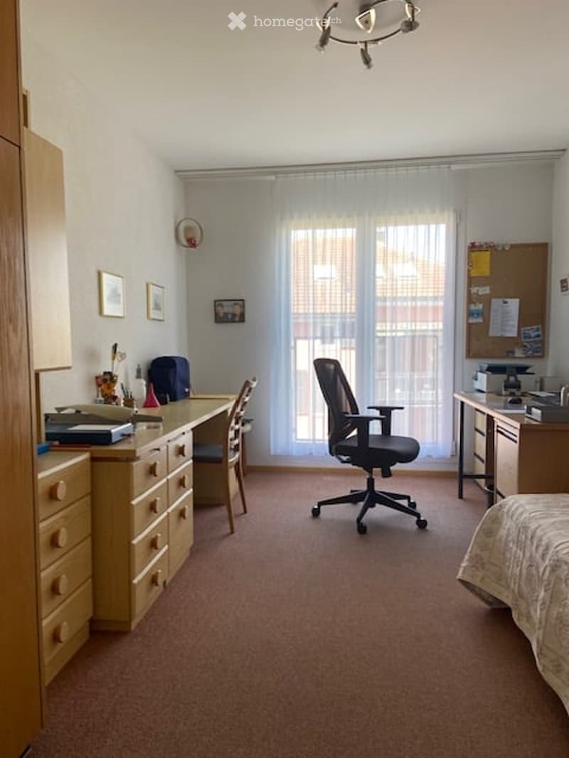 A well-lit bedroom with a desk, office chair, and bed. The room features a large window with white curtains, a wooden cabinet, and several framed pictures on the wall.