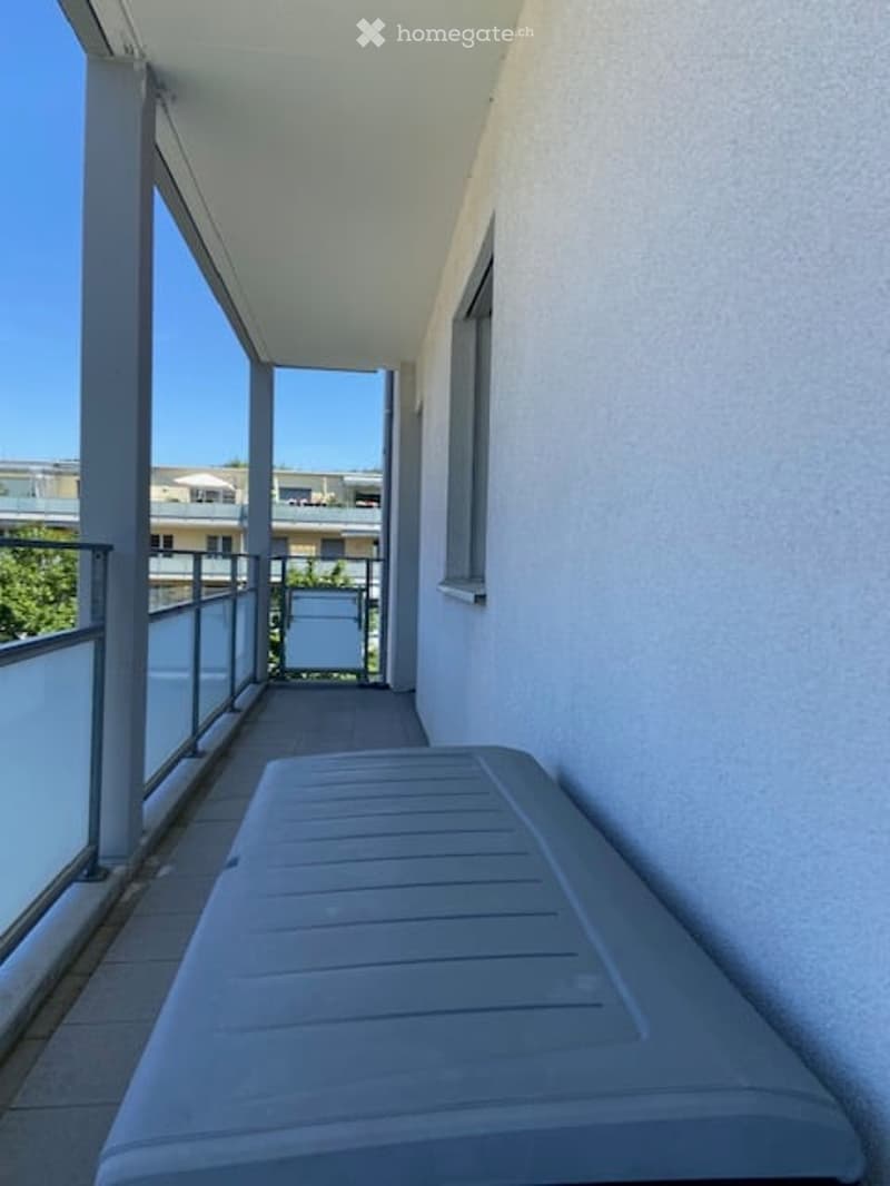 Balcony with white walls, large windows, and a grey storage box on the floor.