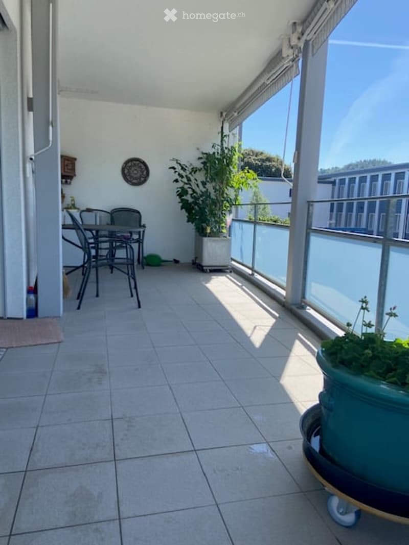 Balcony with tiled floor, glass railing, small table and chairs, potted plants, and a view of the city.