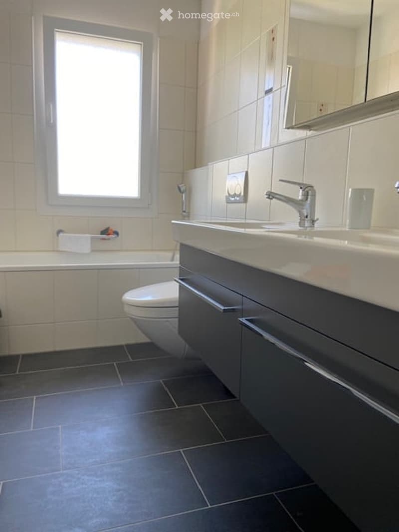 Modern bathroom with dark grey tiles, double sink, large mirror, toilet, bathtub, and a window for natural light.
