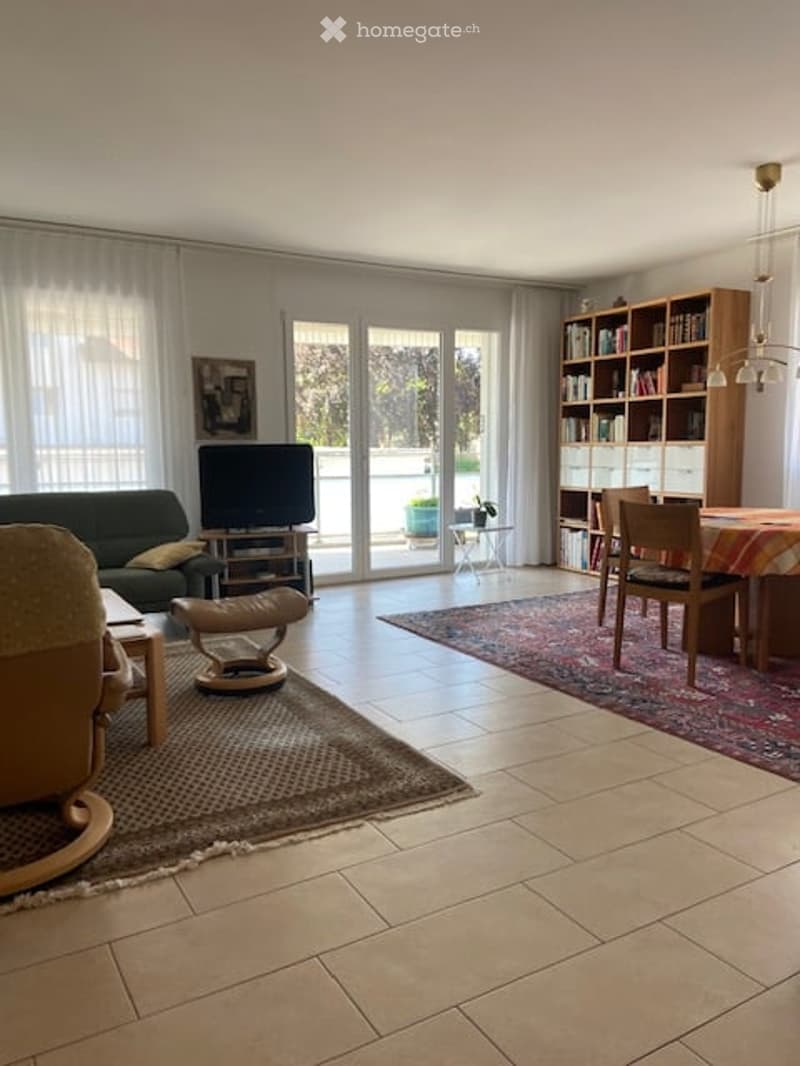 Bright living room with large windows and a view, featuring a green sofa, ottoman, coffee table, dining area with a table and chairs, bookshelf, and a television.