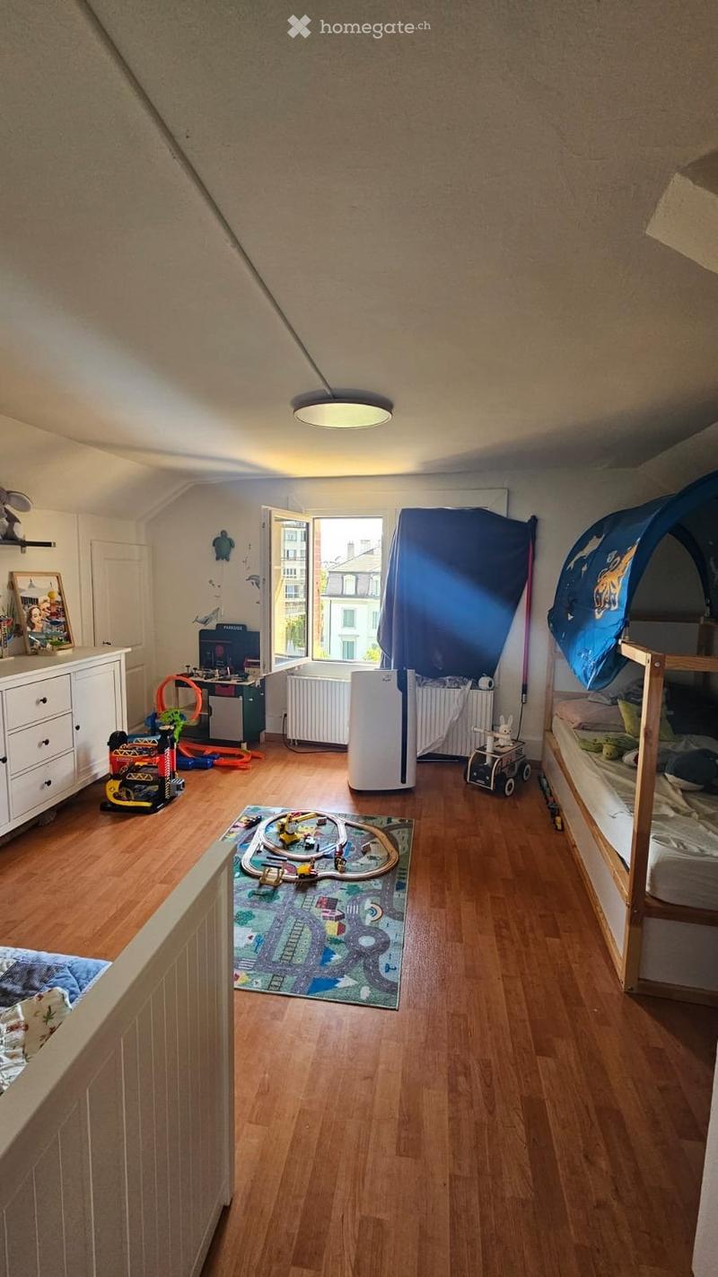 A well-lit children's bedroom with wooden flooring, a bunk bed, a desk with a chair, a toy train set on a rug, a curtained window, a radiator, an air conditioner, and various toys including a toy car and a toy tent.