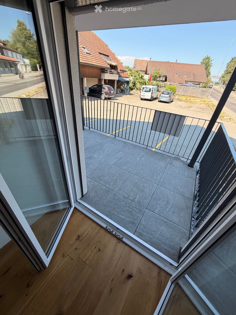 Balcony with modern glass railing, tiled floor, overlooking a parking area with several cars and neighboring houses.