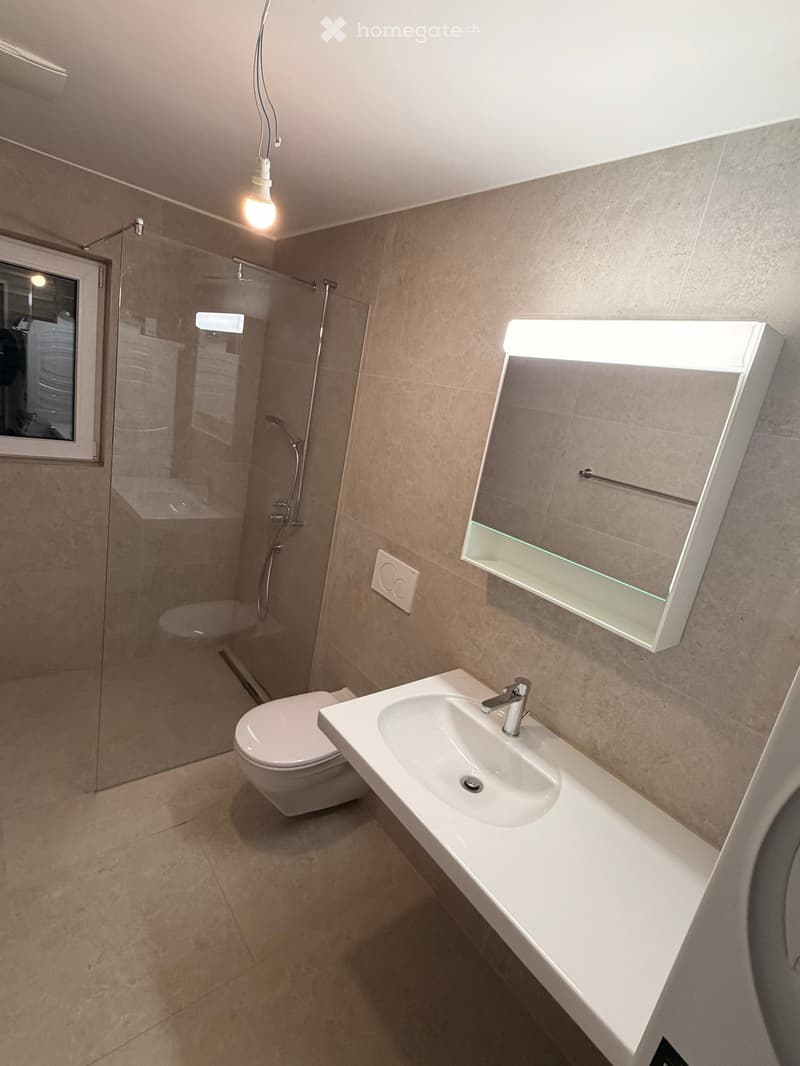 Modern bathroom with a white sink, mirror, toilet, and glass-enclosed shower area. Beige tiled walls and floor, illuminated mirror, and a window providing natural light.