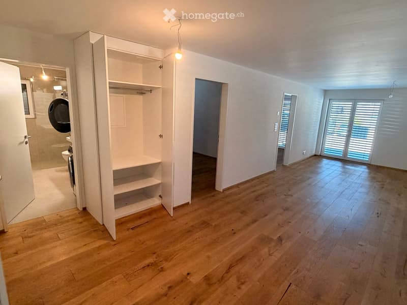 Empty room with wooden flooring, white walls, built-in closet, and large windows with blinds. Adjacent bathroom visible with toilet and shower.