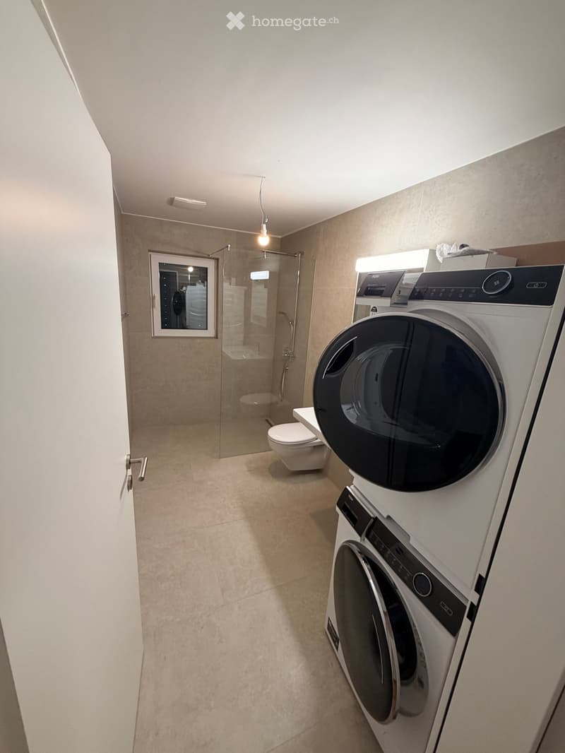 Modern bathroom with a glass-enclosed shower, toilet, and stacked washer/dryer unit. Light-colored tiles and a sleek design.