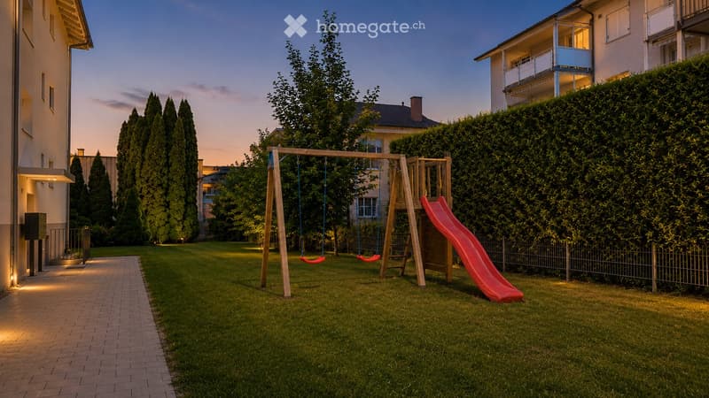 Evening view of a well-maintained garden with a swing set, slide, and surrounding trees, adjacent to a residential building with illuminated balconies.