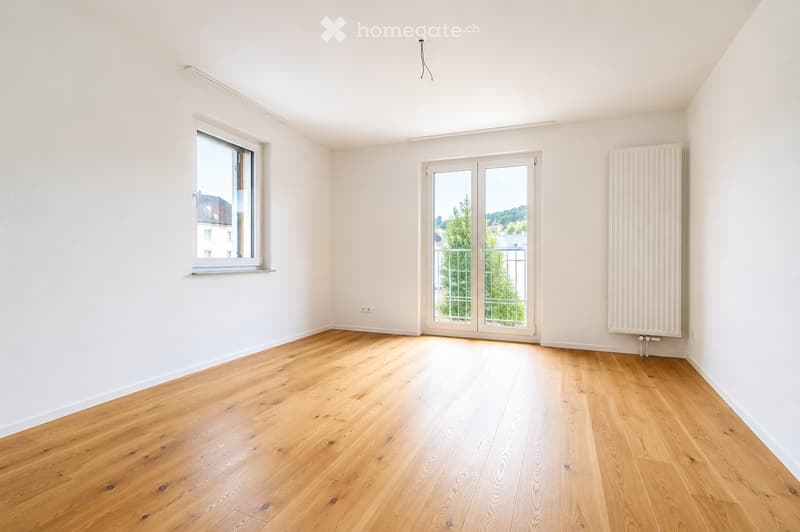 Empty room with light wooden flooring, white walls, large window, and sliding glass door leading to a balcony.