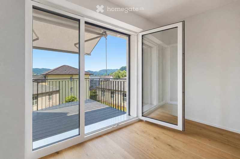 Open sliding glass doors lead to a wooden deck balcony with a metal railing, overlooking a residential area with houses and mountains in the background.