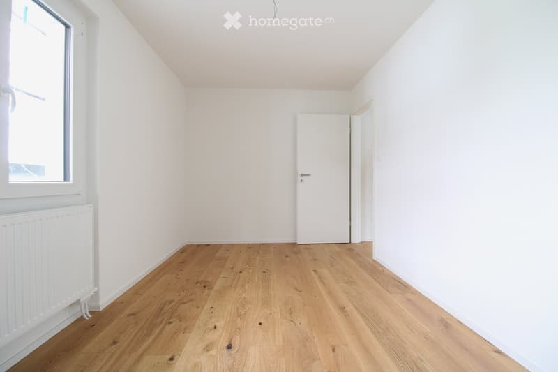 Empty room with light wooden flooring, white walls, a single window, and a white radiator. No furniture or personal items are present.