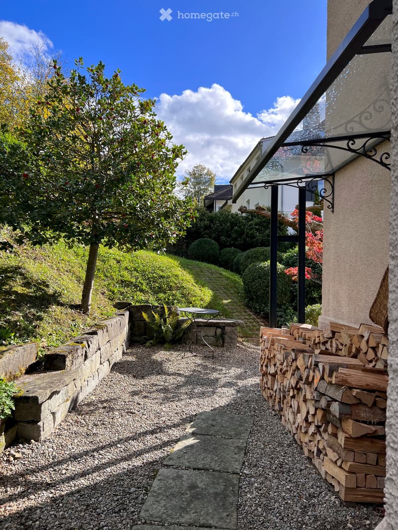 Garden path lined with gravel and stone walls, featuring a small table, neatly stacked firewood, and surrounding greenery.
